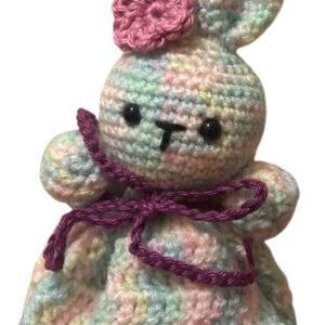 Eine gehäkelte Hasenpuppe mit pastellfarbenem, buntem Garn, schwarzen Knopfaugen, einer rosa gehäkelten Blume an einem Ohr und einer lila Schleife um den Hals.